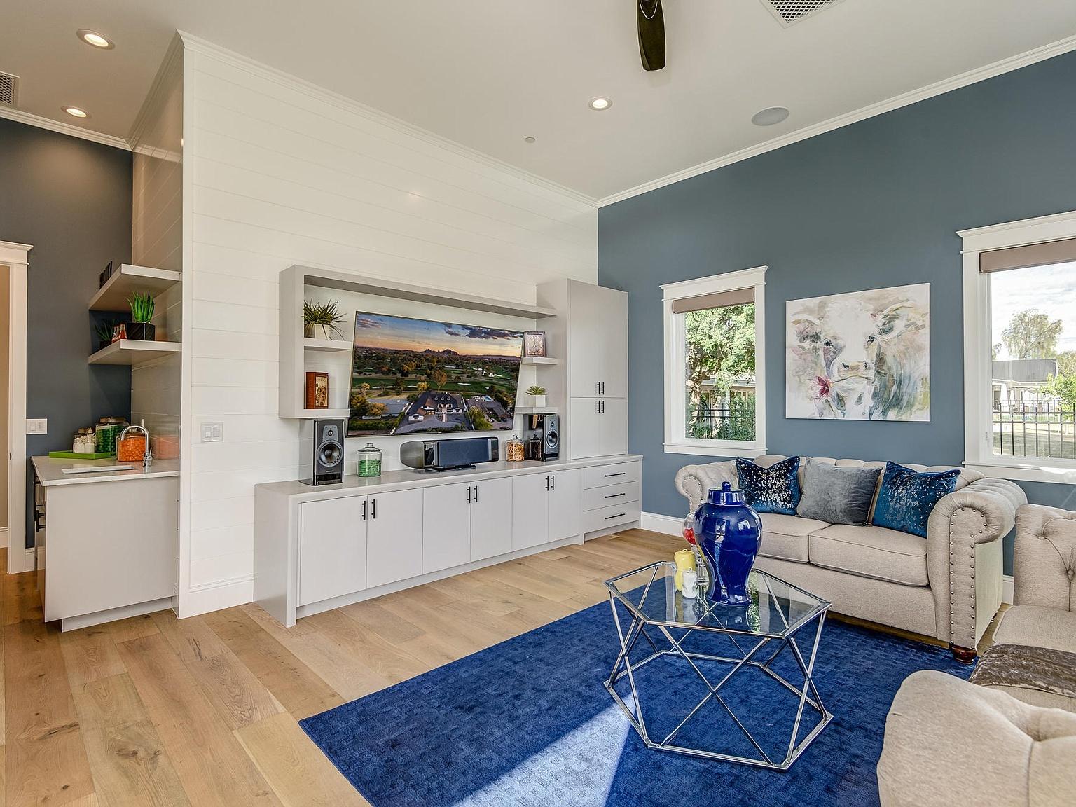 Bright contemporary living room with blue accent walls, a built-in white media cabinet and TV, light beige tufted sofas with blue pillows, and a glass coffee table on a deep blue area rug.
