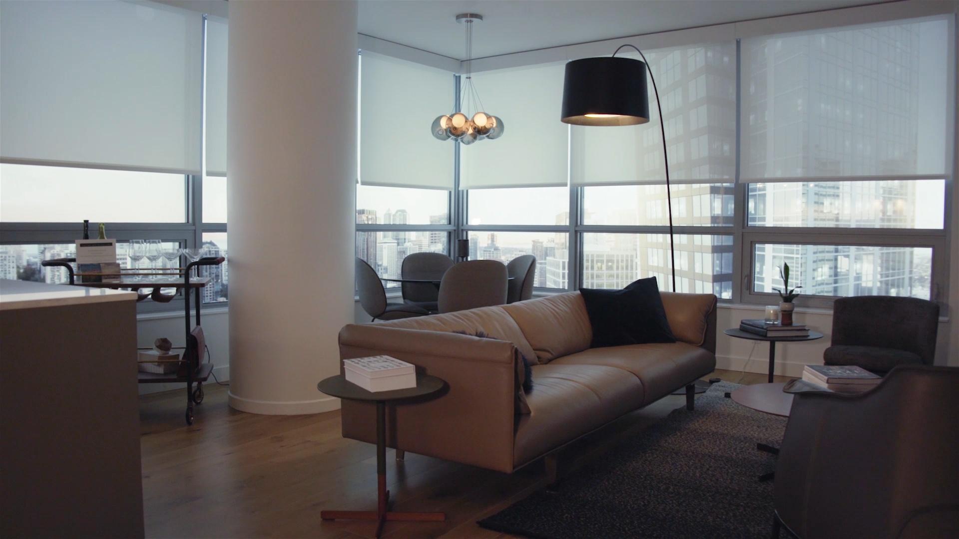 Modern corner apartment living and dining area with floor-to-ceiling windows and roller shades, a gray sectional sofa, round dining table with four chairs, and a large white column on light wood floors overlooking a city skyline.
