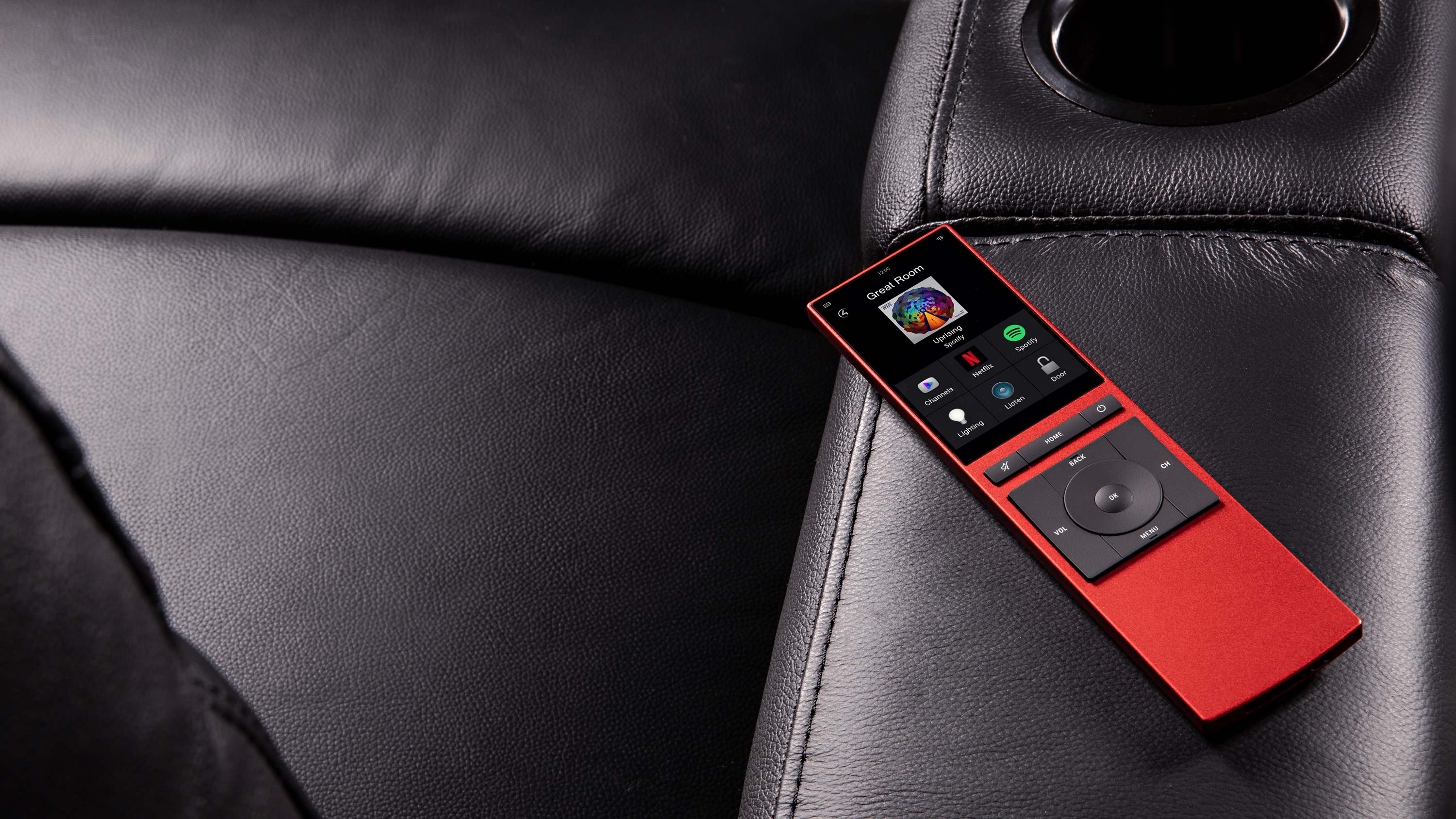 Modern home theater with a red smart remote resting on a black leather surface, showing a touchscreen interface for controlling lighting, streaming apps, and media