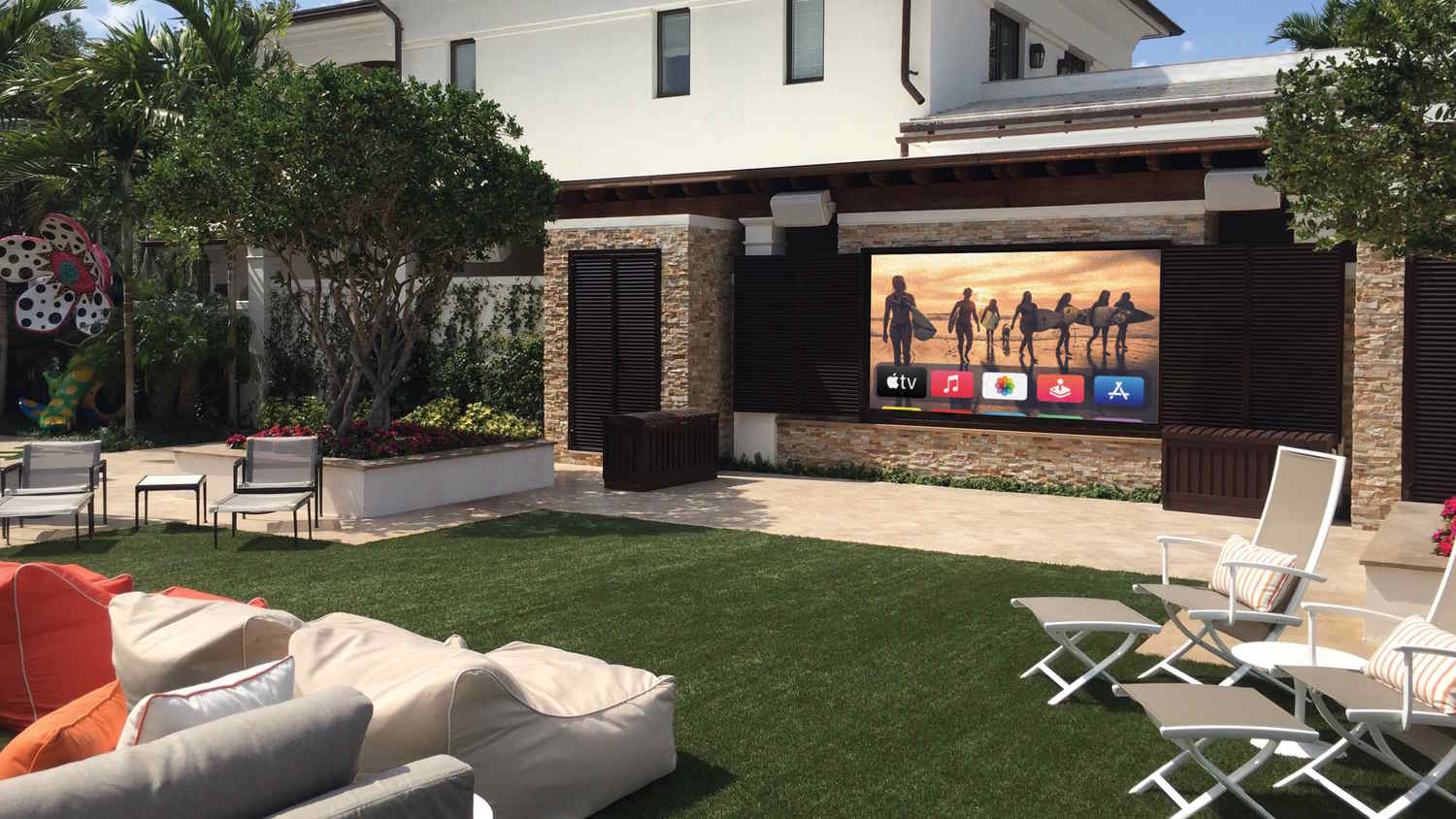 Luxurious outdoor lounge with a large TV, cushioned seating, and a landscaped garden backdrop.
