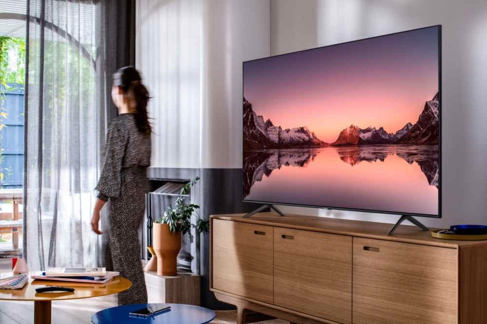 Modern living room with a large TV on a wooden console, a round coffee table, and someone standing by sheer curtains with greenery outside.