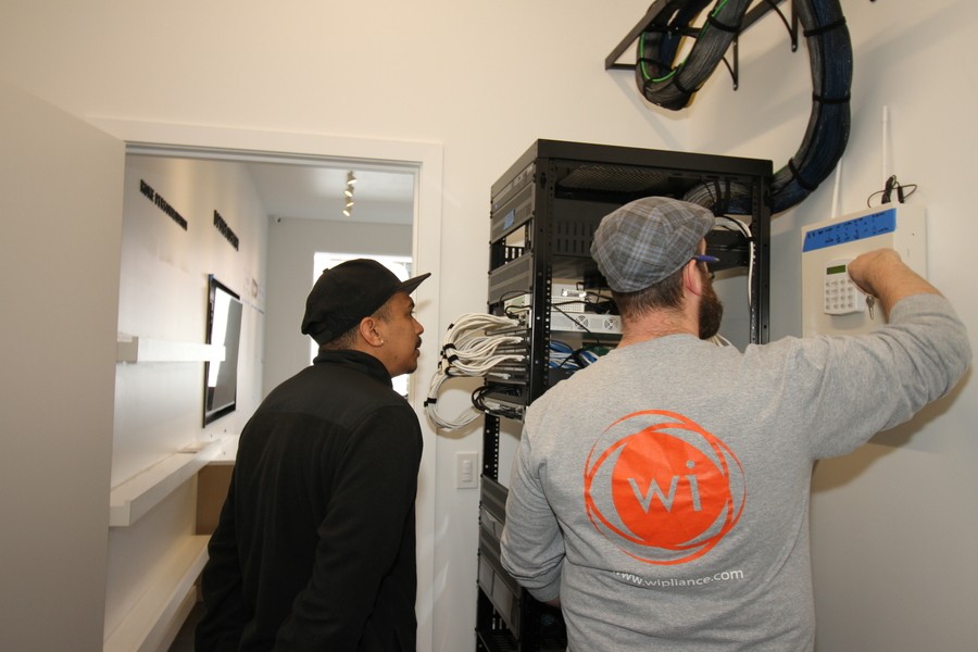 two Wipliance technicians work on an AV rack and keypad in a client’s hallway