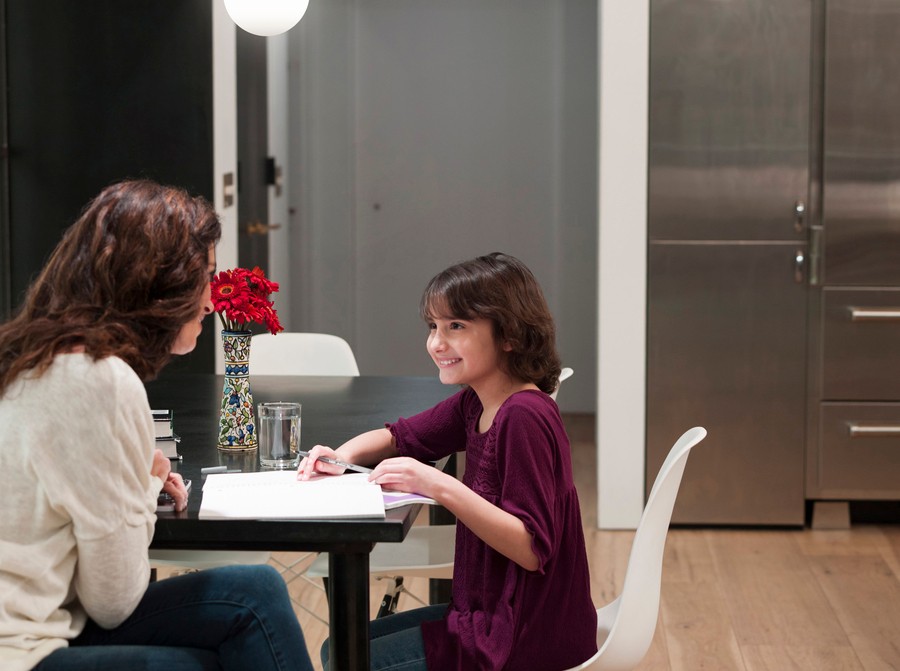 Mother and daughter sitting at a kitchen table, doing homework together under bright, modern lighting, with flowers in a vase nearby. 