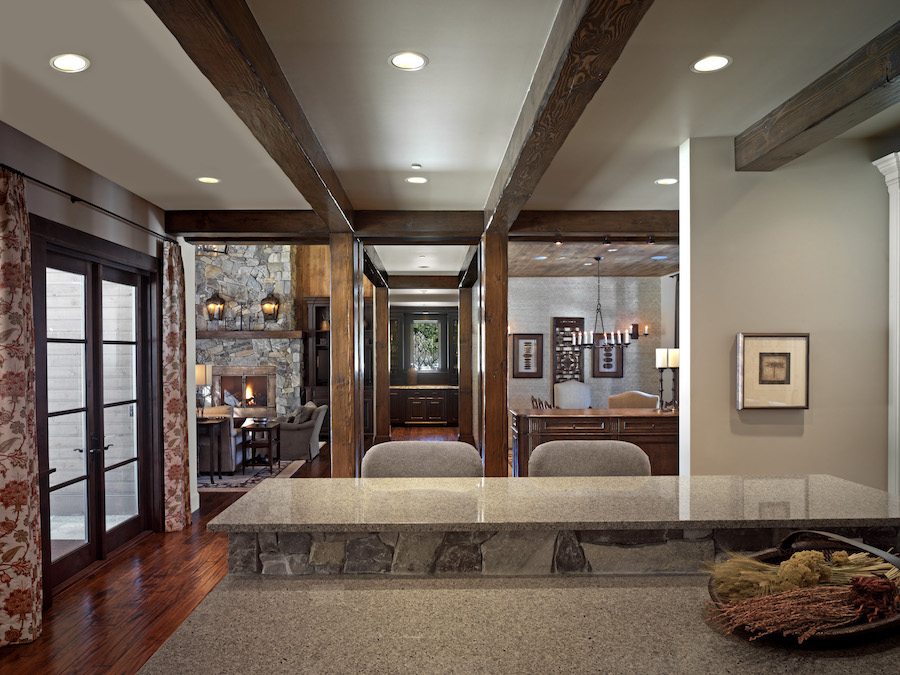 Open-concept rustic living space viewed from behind a stone-front kitchen island, featuring exposed wood beams on the ceiling, French doors with patterned curtains on the left, a stone fireplace seating area in the background