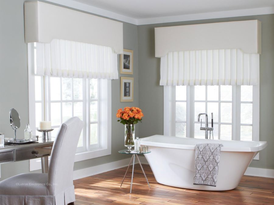 Bright, elegant bathroom with a freestanding white bathtub in front of two large windows with soft valances, a gray vanity and upholstered chair on the left, and a small glass table holding orange flowers and bath items beside the tub.