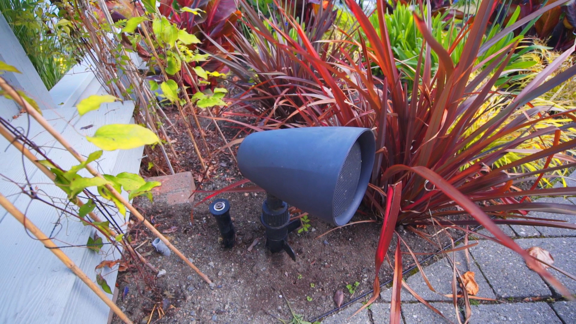 Outdoor bullet-style landscape speaker staked in a garden bed among colorful plants next to a walkway.