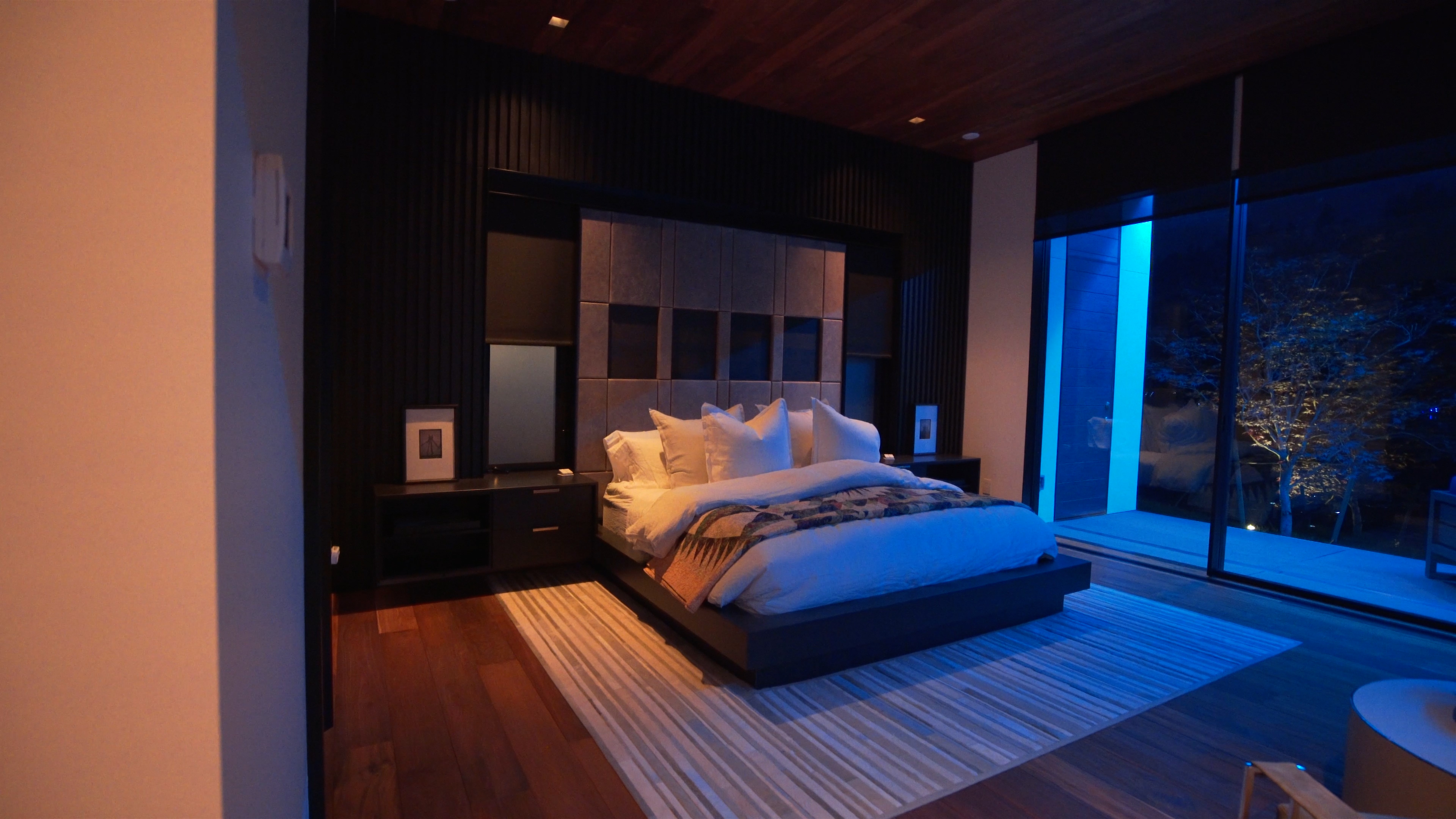 A cozy bedroom featuring blue ambient lighting, creating a relaxing atmosphere with floor-to-ceiling windows.