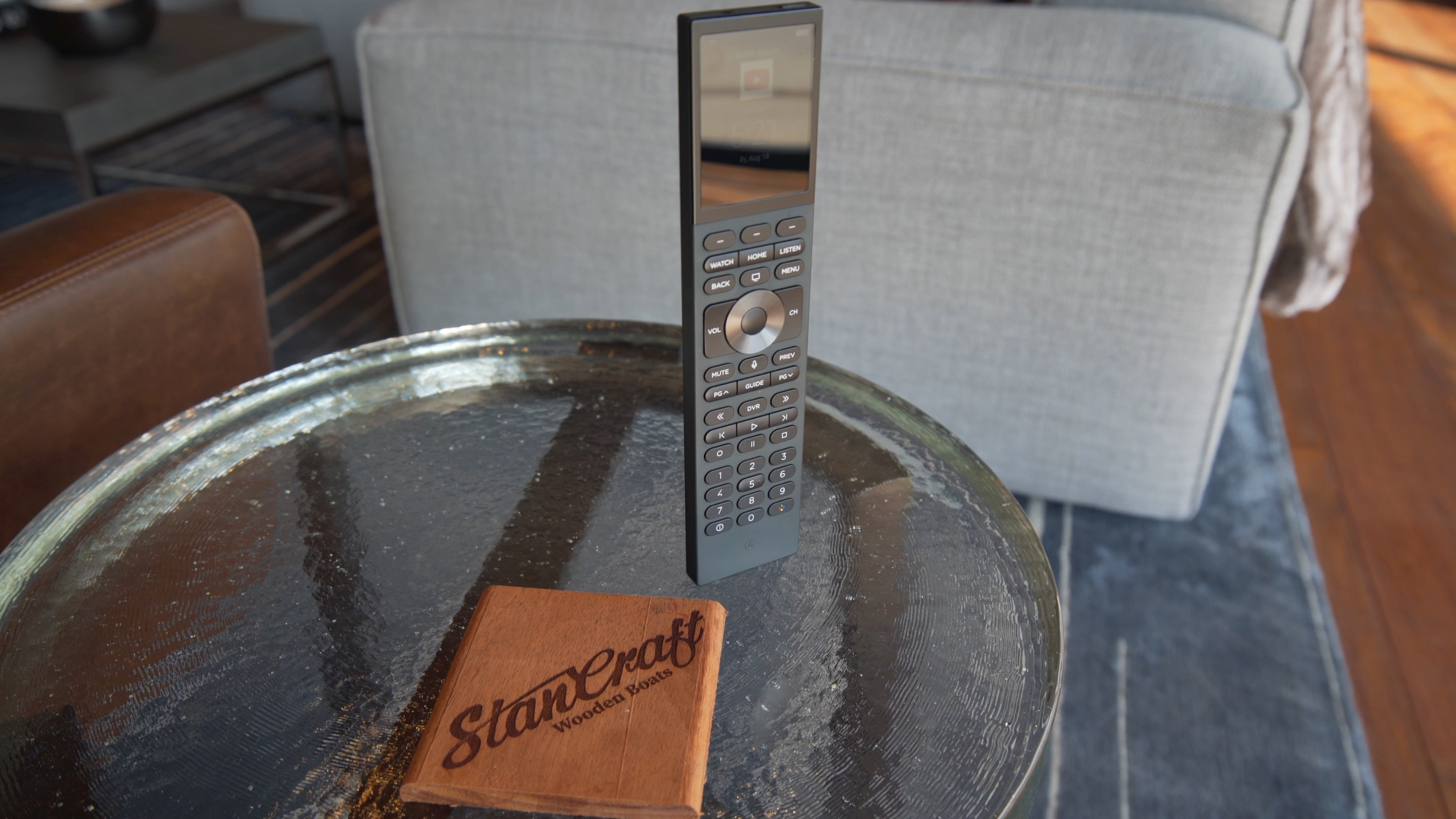 n a modern living space, a sleek smart home remote rests on a glass table with a wooden coaster.