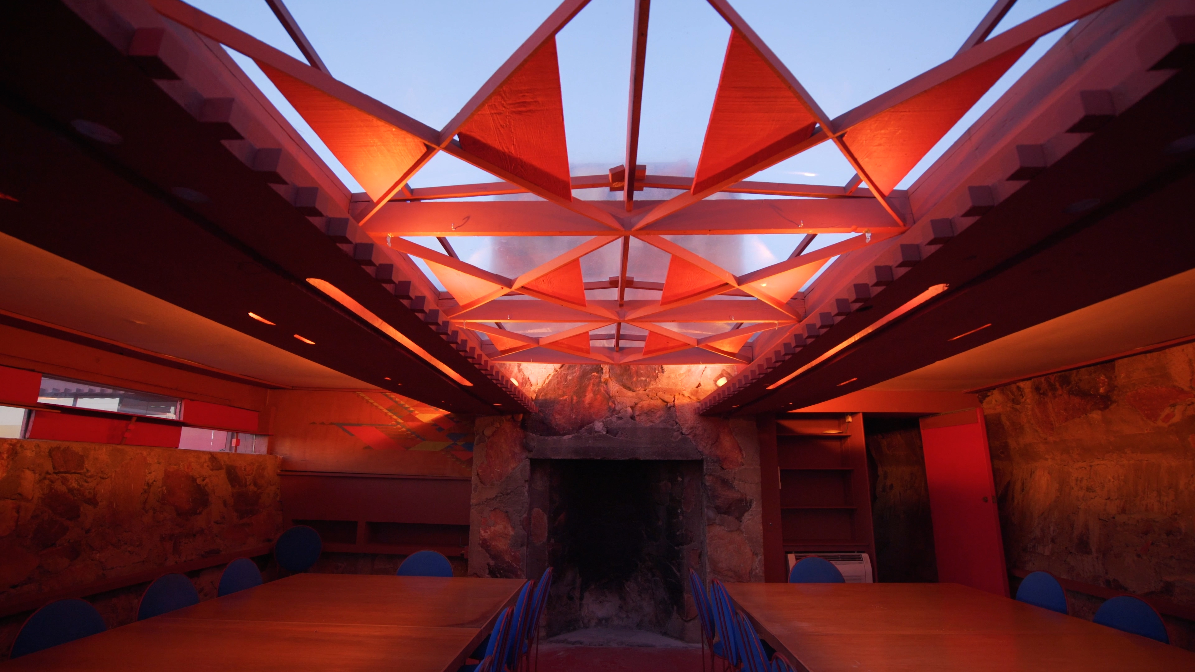 Low-ceilinged boardroom at Taliesin West lit by red‑hued LED lighting under geometric skylight and around stone fireplace.