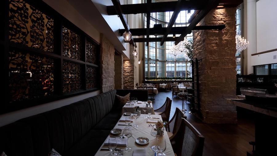 Elegant restaurant dining room with layered pendant lighting, stone columns, and a sunlit atrium skylight.