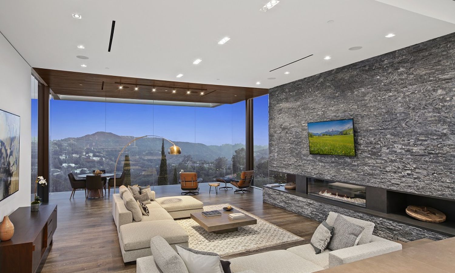 Open-concept luxury living room with a stone fireplace wall, wall-mounted TV, and floor-to-ceiling windows framing mountain views