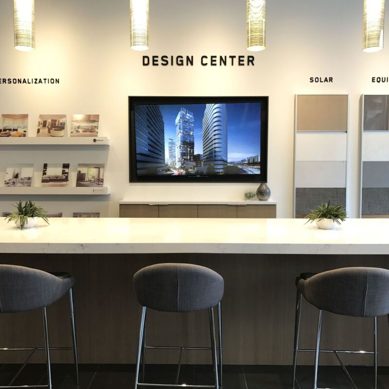 &ldquo;Design Center&rdquo; showroom with a long white counter and three gray barstools facing a wall-mounted screen, surrounded by sample boards, brochures, and pendant lights.