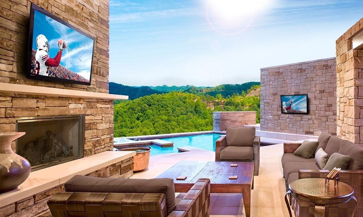 Covered outdoor living area with a TV above a stone fireplace, lounge seating, and a pool overlooking green hills