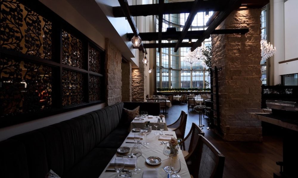 Elegant restaurant dining room with layered pendant lighting, stone columns, and a sunlit atrium skylight.
