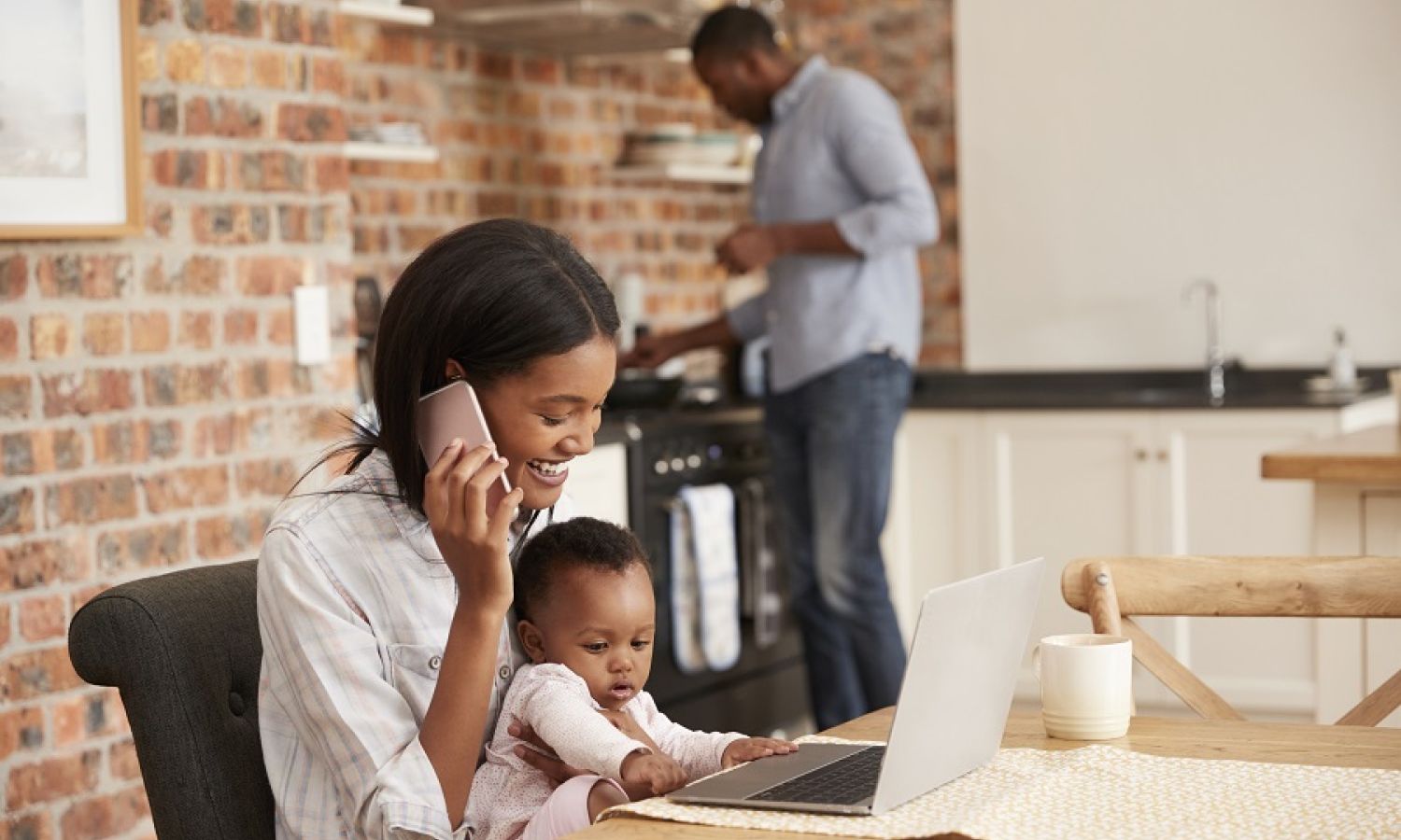 Parent working on a laptop at the kitchen table with a toddler while another adult stands in the kitchen
